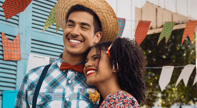 Casal jovem de homem e mulher com trajes caipiras e bandeirolas de festa junina ao fundo, em artigo sobre charraiá de fraldas