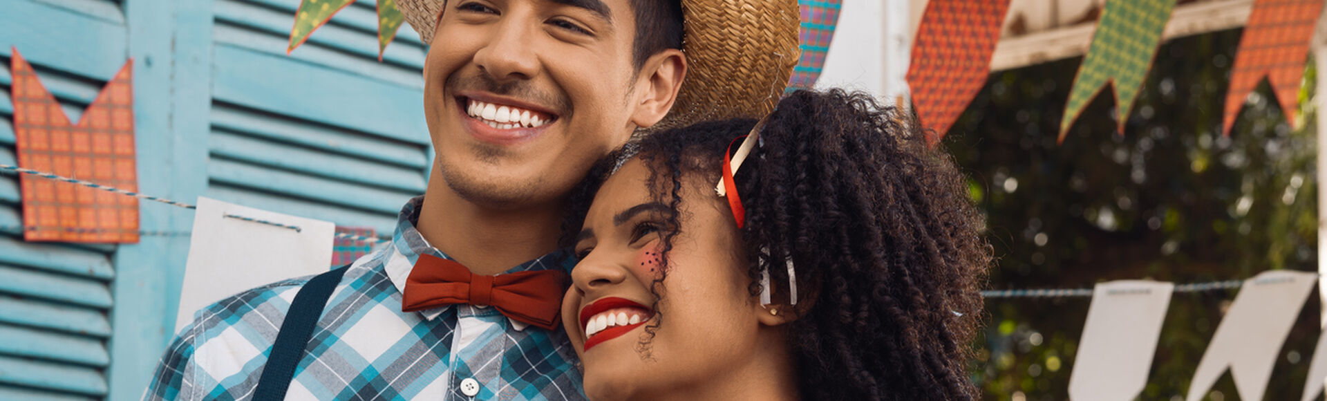 Casal jovem de homem e mulher com trajes caipiras e bandeirolas de festa junina ao fundo, em artigo sobre charraiá de fraldas