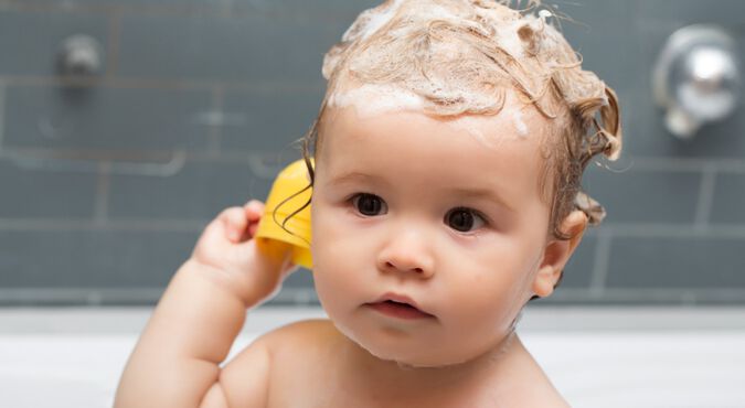 Bebê branco tomando banho de banheira com espuma de shampoo de bebê em seus cabelos castanhos. Ele aparece do ombro para cima