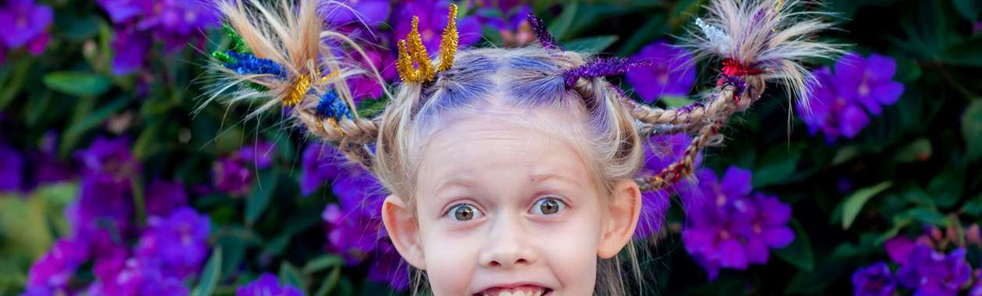 Garota feliz, com olhos arregalados e penteado inspirado nas flores roxas que estão no arbusto atrás dela.