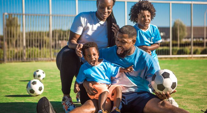 Imagem de uma família jogando futebol em um campo ao ar livre. A família inclui um pai, uma mãe e dois filhos pequenos, todos vestidos com camisetas esportivas azuis.