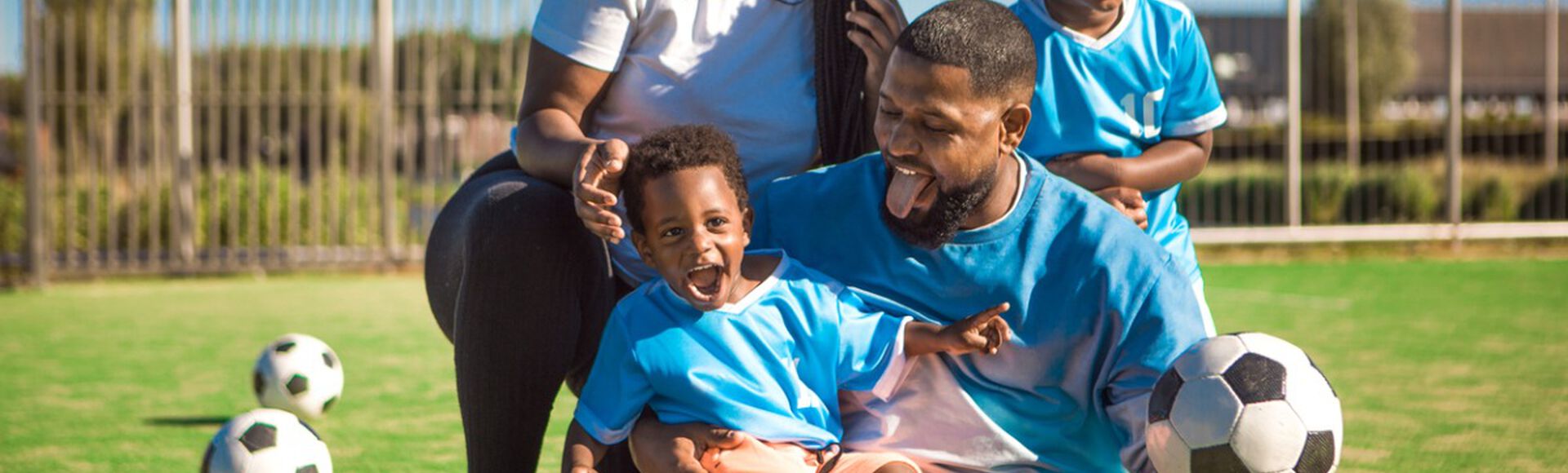 Imagem de uma família jogando futebol em um campo ao ar livre. A família inclui um pai, uma mãe e dois filhos pequenos, todos vestidos com camisetas esportivas azuis.