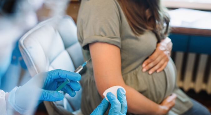 Mulher branca gestante recebendo vacina no consultório médico. Rosto da grávida e de quem aplica a vacina não aparecem.