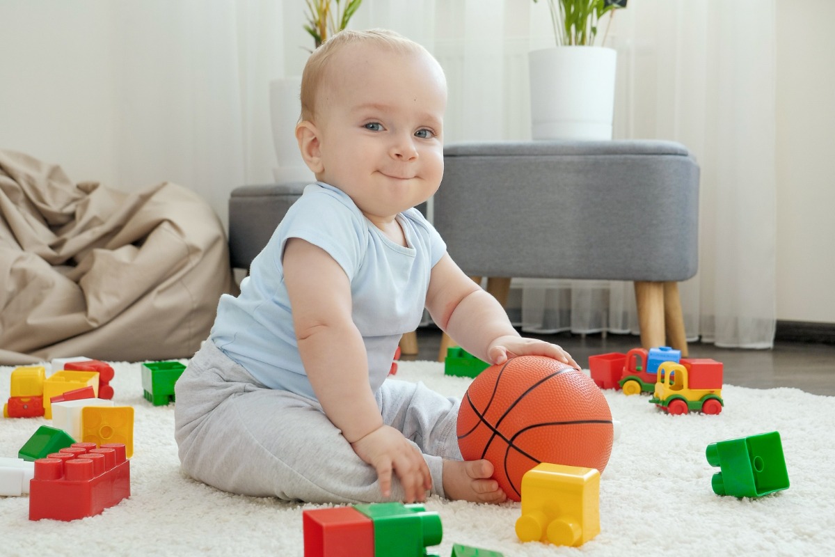 Bebê branco brinca no chão com peças de montar e bola de basquete, em artigo sobre visão do bebê. Está sobre tapete branco 
