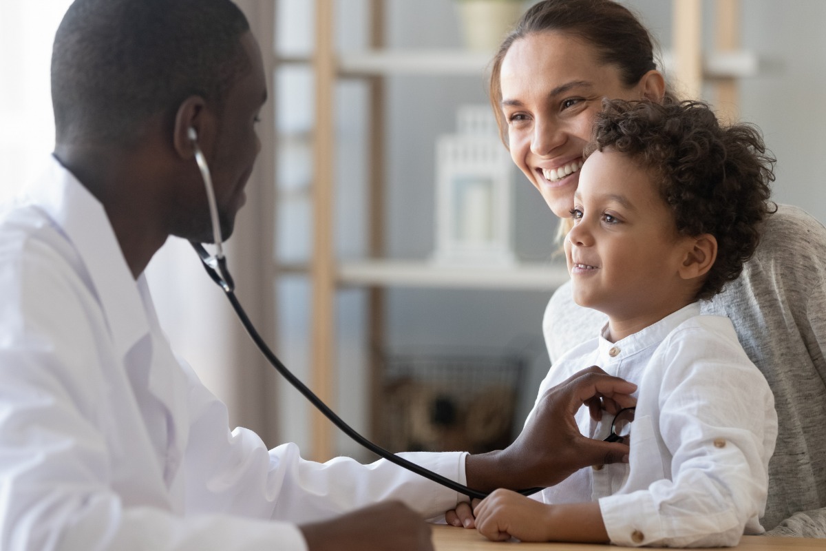 Imagem de um médico examinando um menino com um estetoscópio enquanto a mãe do menino observa com um sorriso.