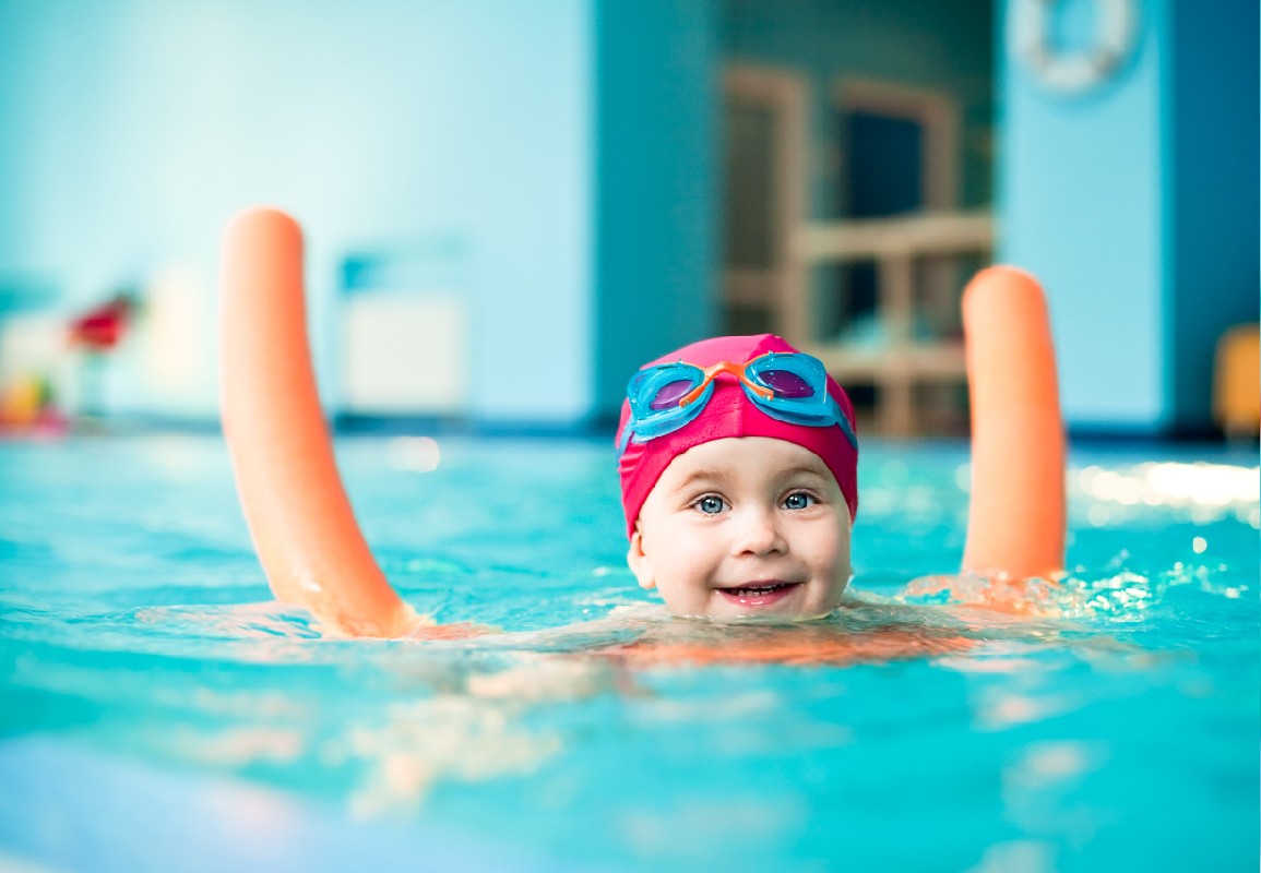 criança nadando em uma piscina com uma touca de natação vermelha e óculos de natação azuis. A criança está usando dois flutuadores de espuma laranja e sorrindo para a câmera.