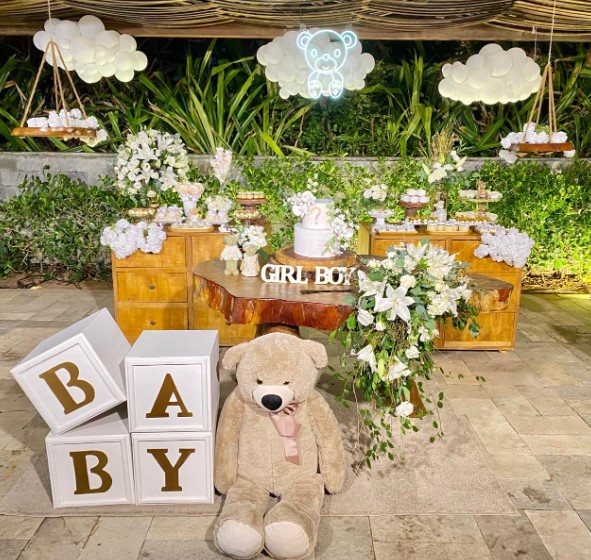 Decoração bem naturalista de chá revelação, com flores brancas, ursos e objetos em tom madeira e branco, além de plantas ao fundo.