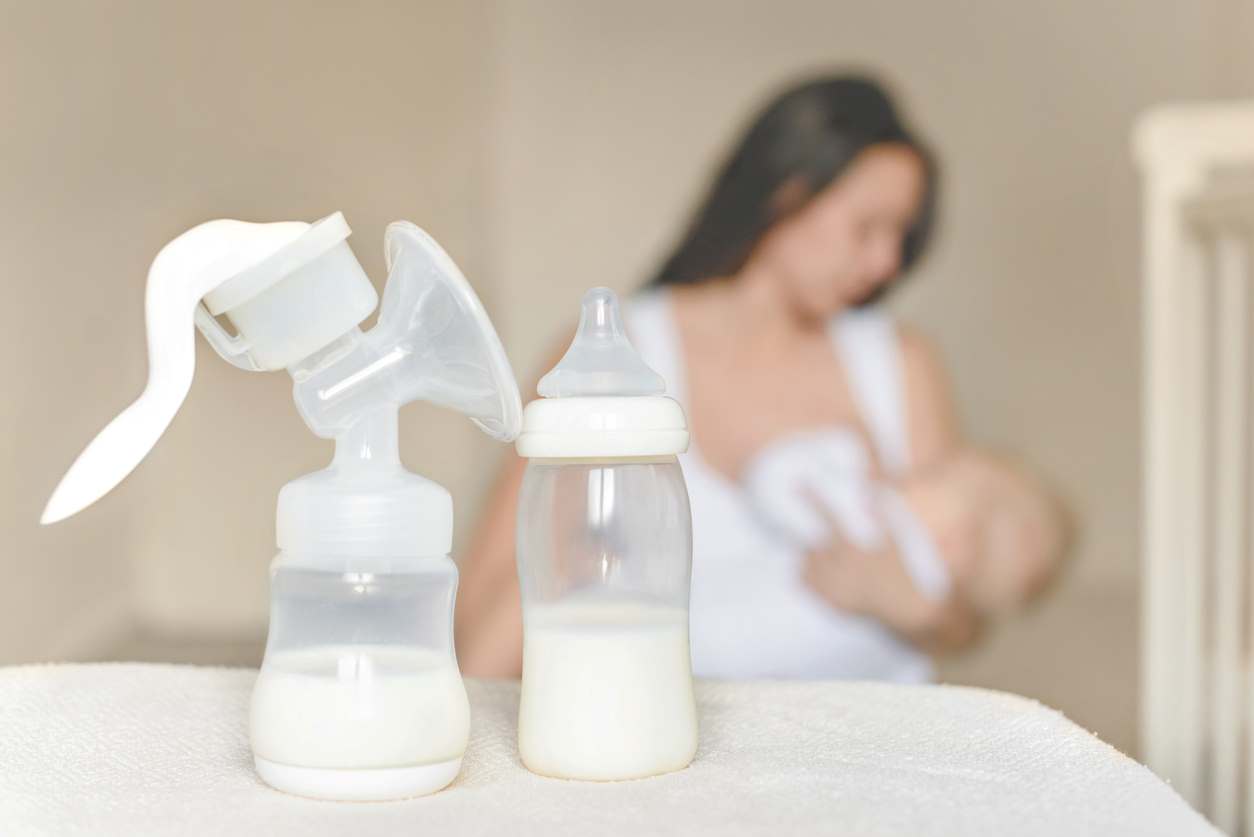 Bomba manual de extração de leite materno ao lado de mamadeira com leite, com mãe amamentando ao fundo.