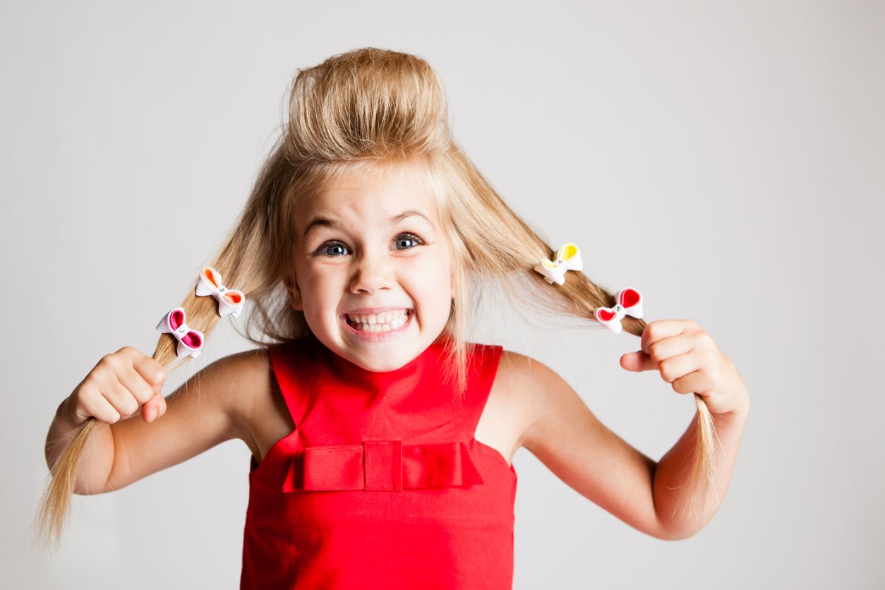 Menina segurando mechas do cabelo enfeitadas com lacinhos e fazendo careta divertida para a foto.