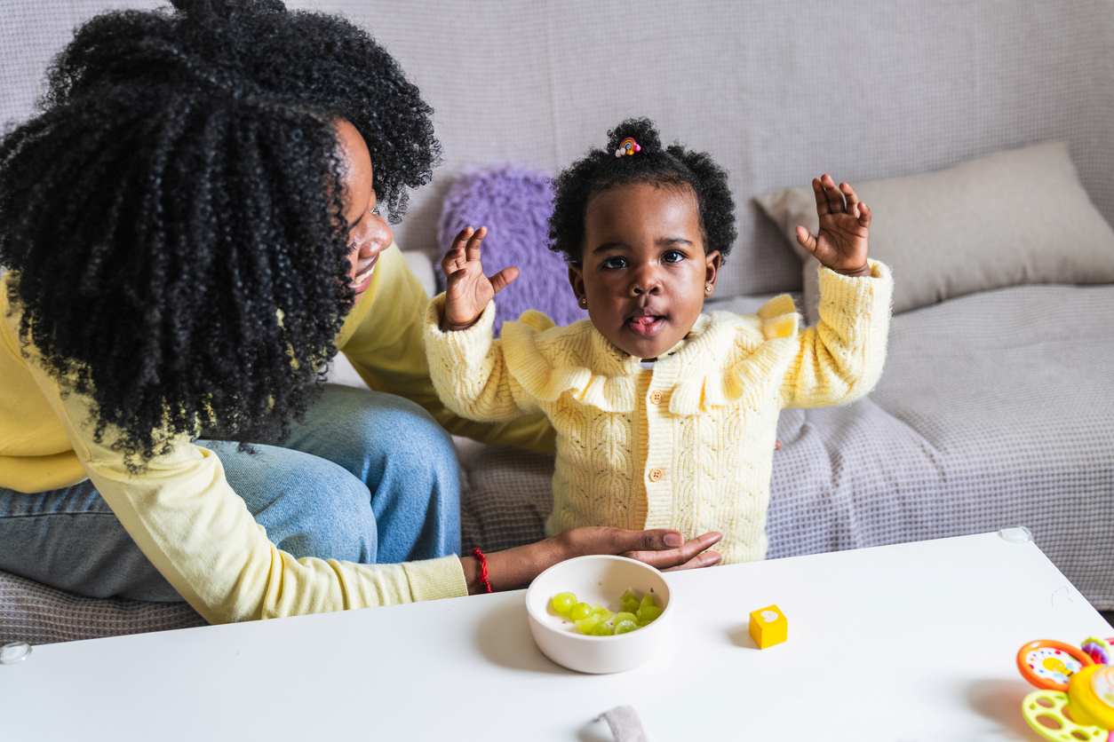 Bebê com presilha divertida no cabelo crespo curto, ao lado da mãe, enquanto come uva na sala de casa