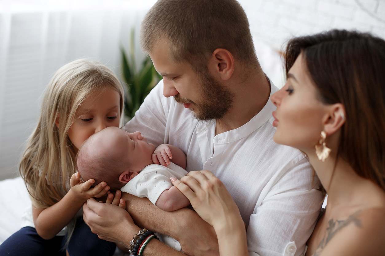 Família reunida. O bebê dorme no colo do pai, de um lado está a mãe, e do outro a irmãzinha, que está beijando a cabeça do irmão.