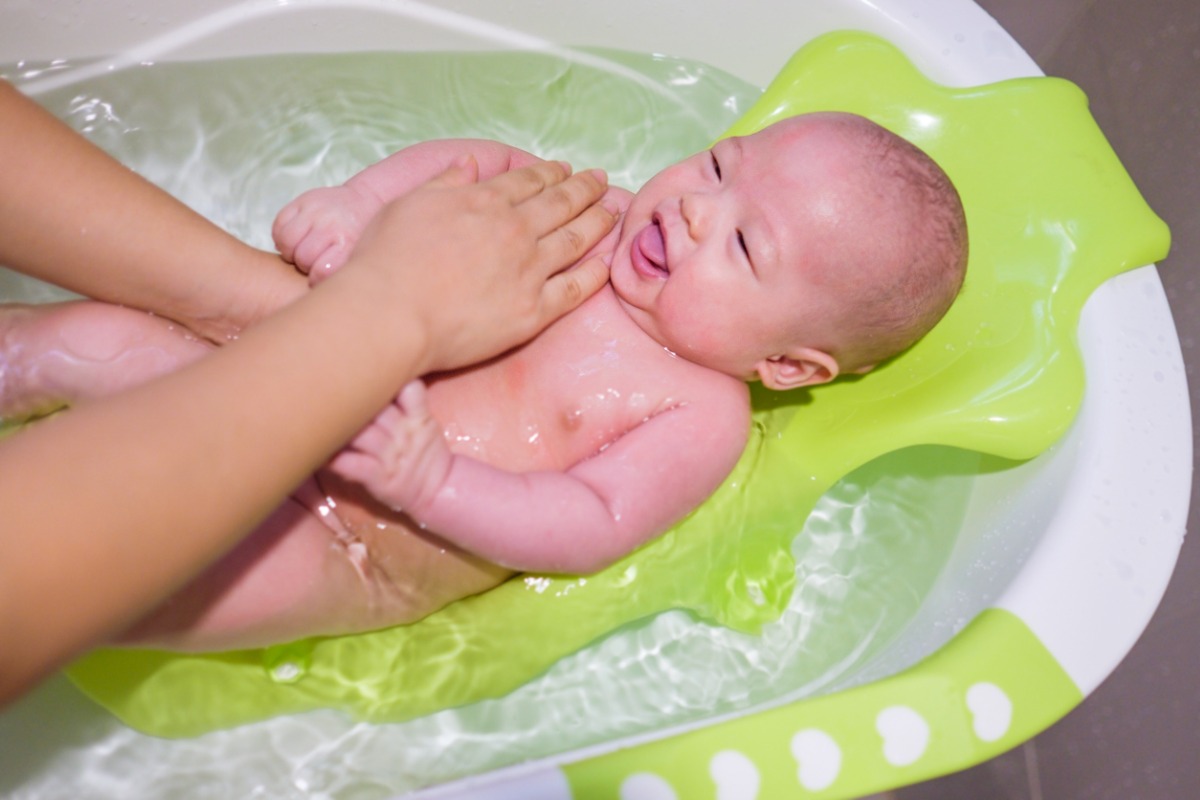 Bebê asiático feliz deitado em banheira de plástico branca com verde. Ele sorri enquanto mãos da cuidadora o acomodam 