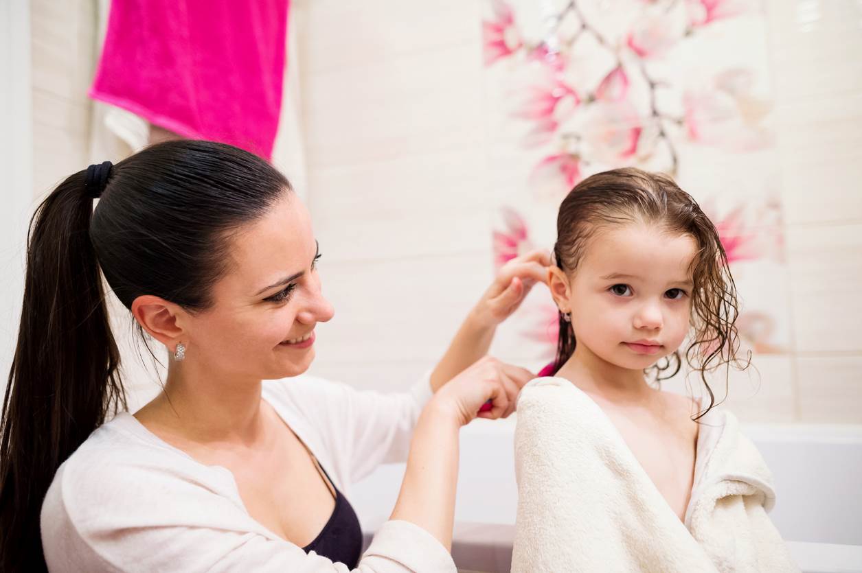 Mãe aplicando creme de pentear infantil no cabelo da filha após o banho.