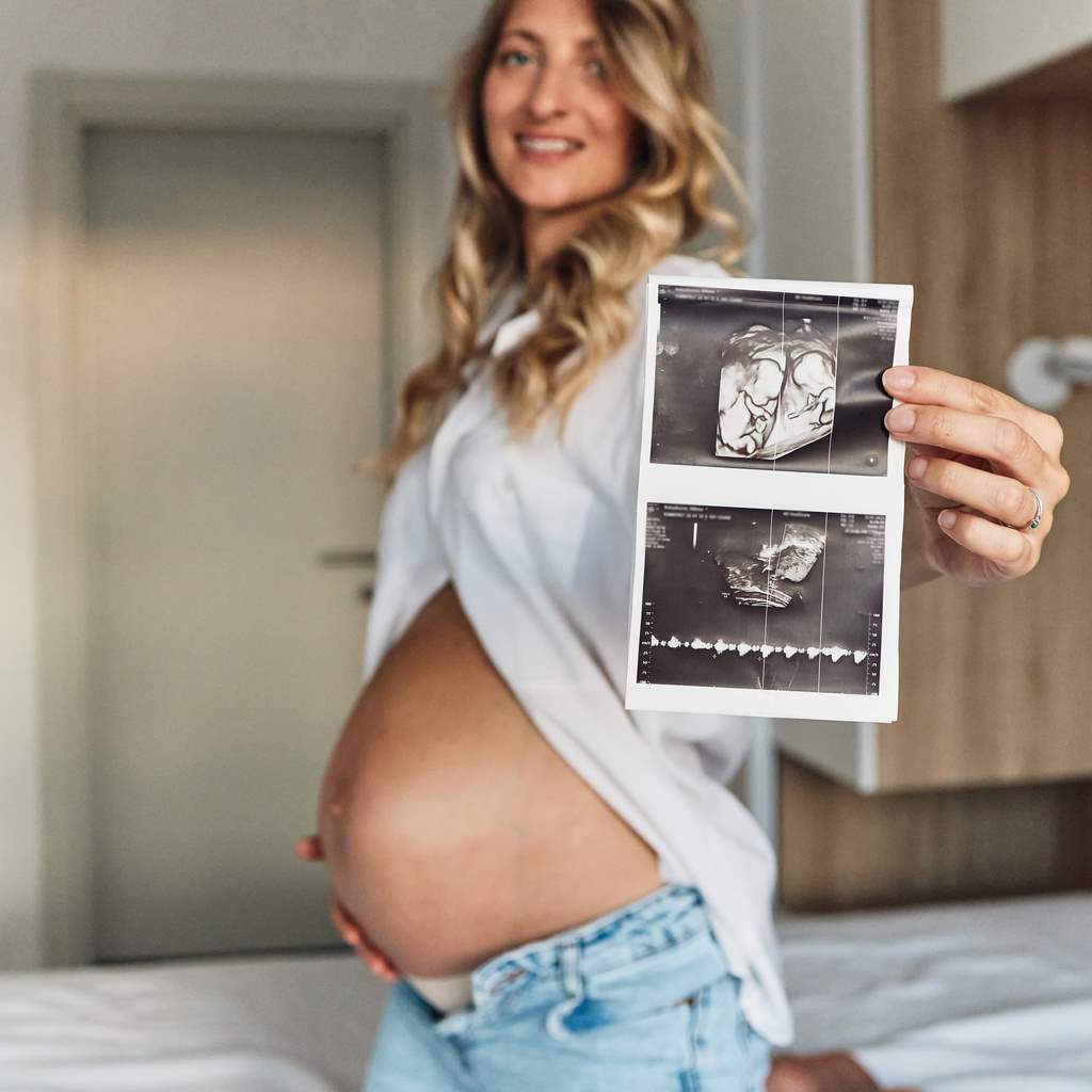 Mulher grávida sorridente segurando duas imagens de ultrassom enquanto exibe sua barriga. 