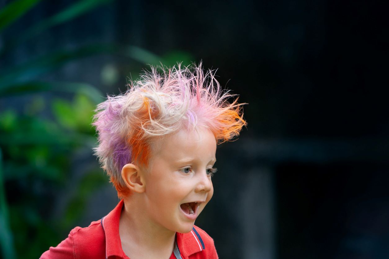 Detalhe de menino com cabelo de três cores, ruivo, loiro e lilás, parece estar correndo feliz (na foto vemos só cabeça e busto).