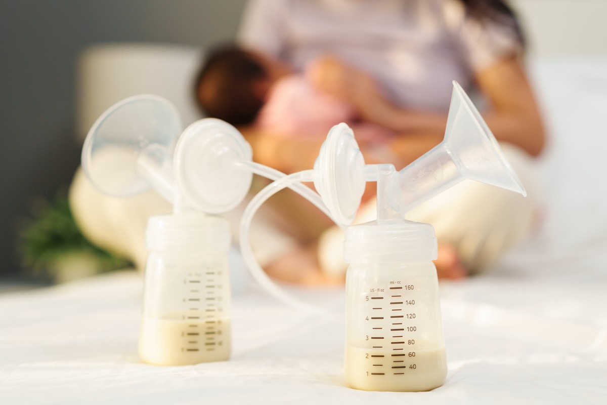 duas mamadeiras com bombas de extração de leite materno em foco, enquanto ao fundo, desfocado, uma mãe amamenta seu bebê. 