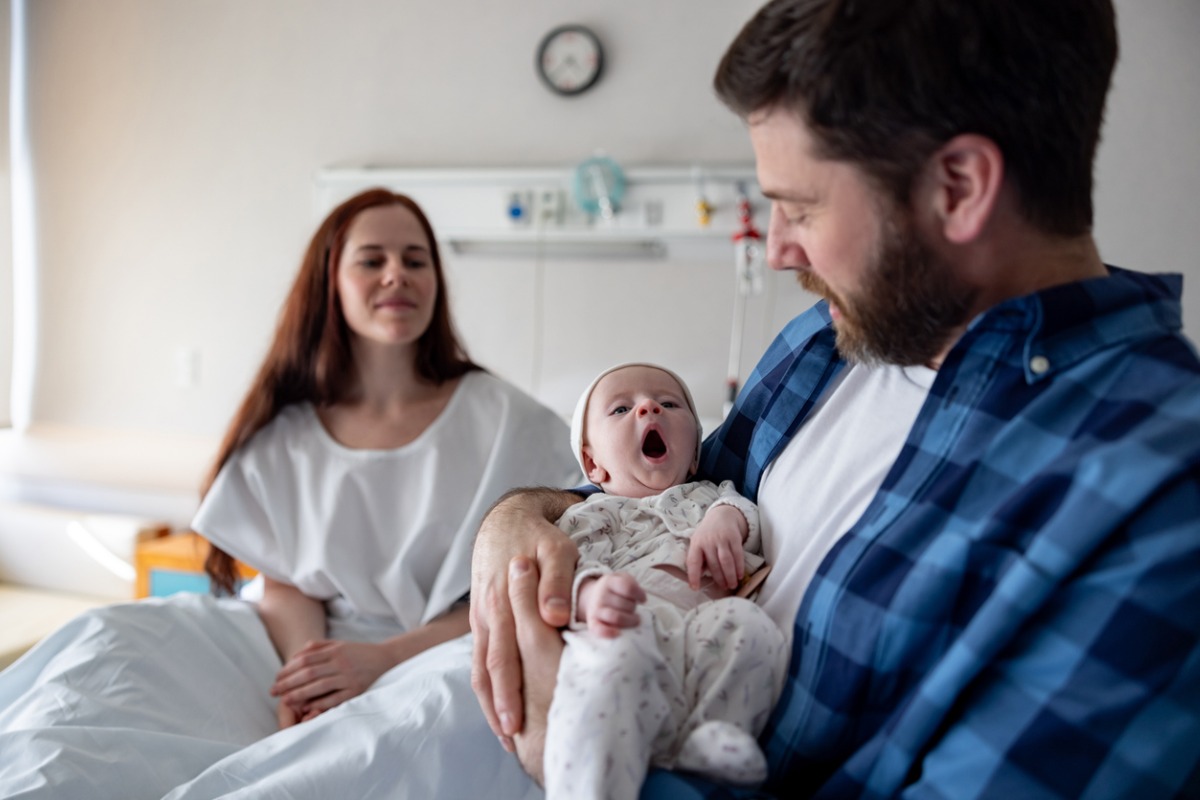 família no hospital, o pai está segurando o bebê recém-nascido que está bocejando, enquanto a mãe, sentada na cama do hospital, observa com um sorriso