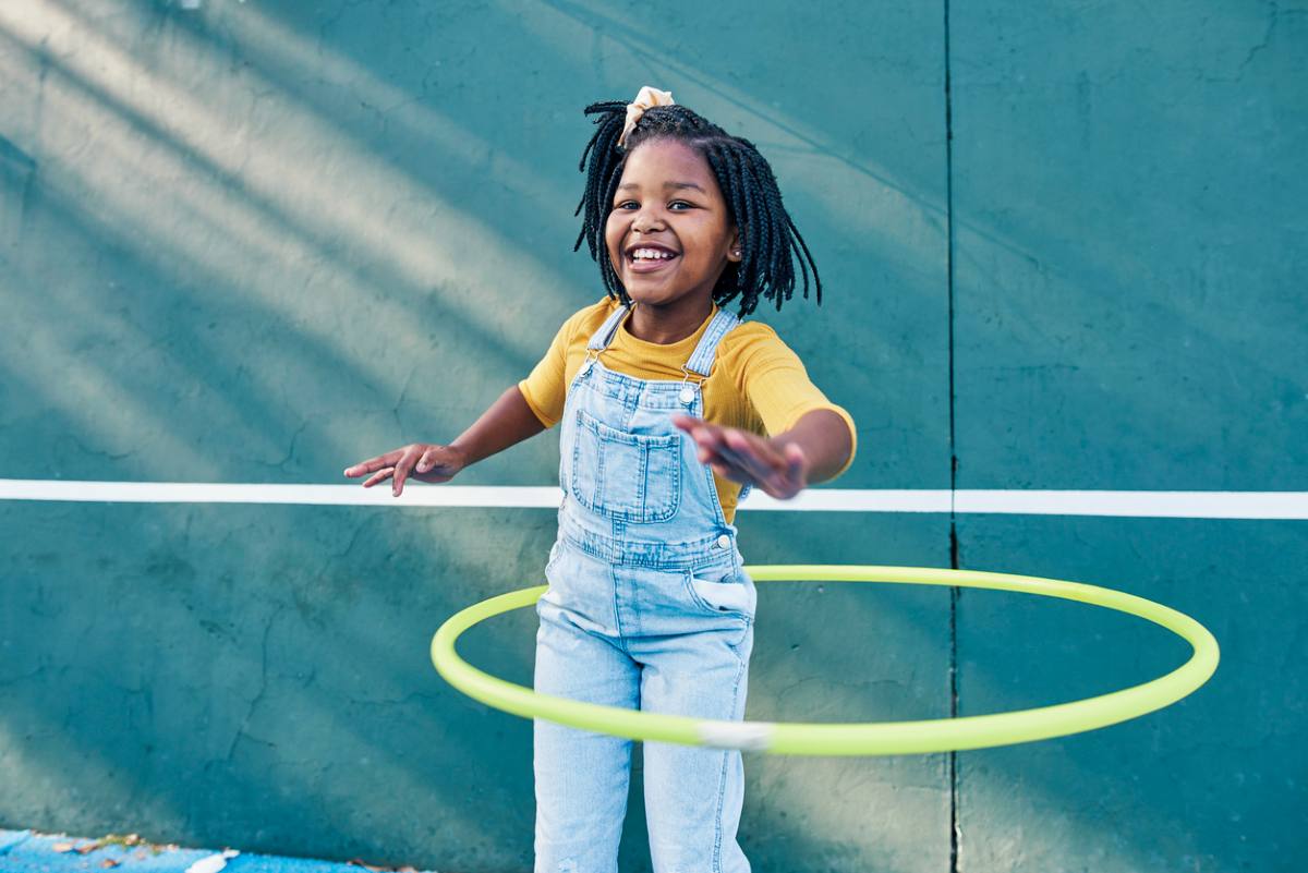 menina brincando com um bambolê verde ao ar livre. A menina está sorrindo e usando um macacão jeans sobre uma camiseta amarela de mangas compridas.