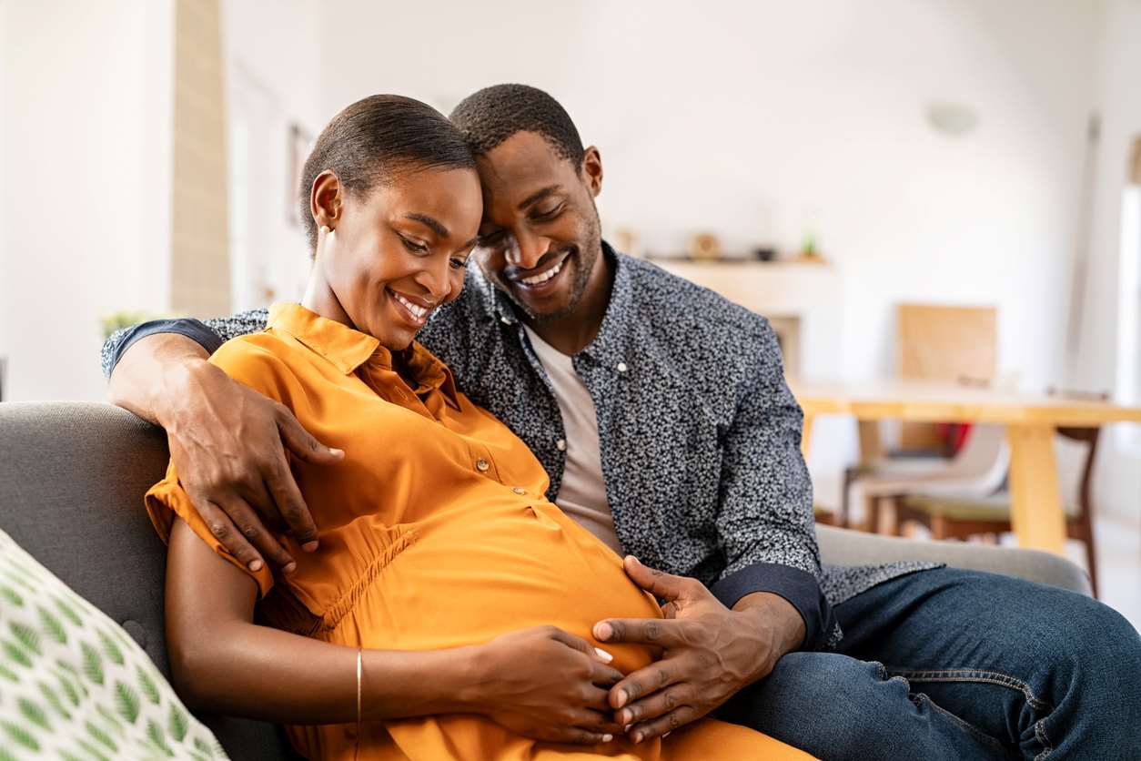 Casal sentado em sofá. A mulher está grávida, o homem a abraça. Os dois olham para a barriga e a tocam gentilmente.