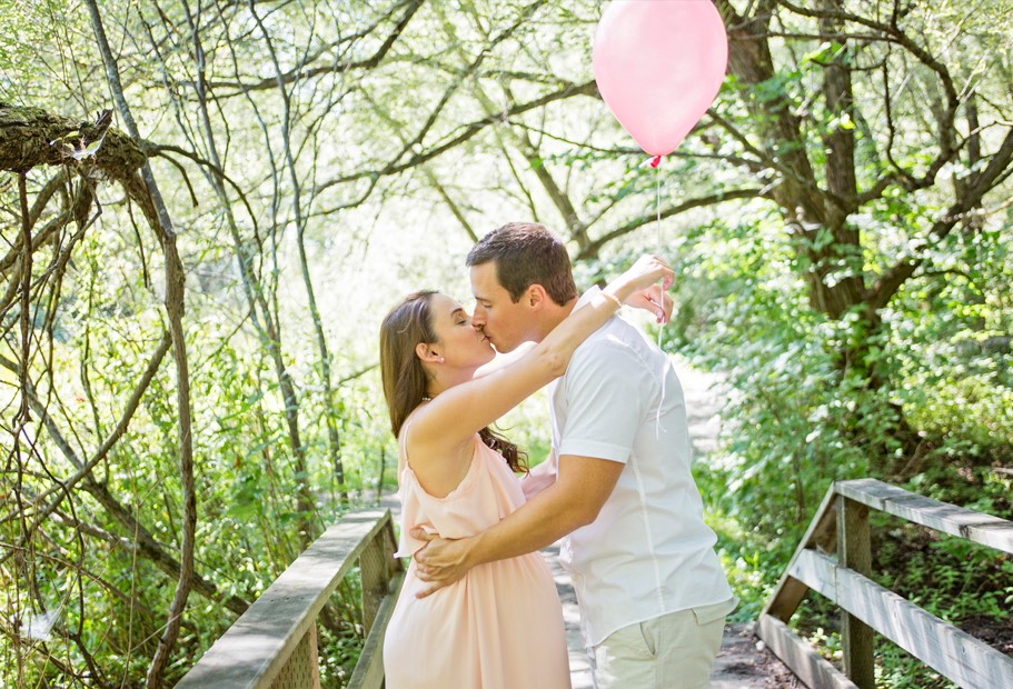 Casal se beijando em uma ponte de madeira, em meio á natureza. A mulher esta grávida e segura um balão rosa, enquanto o marido a segura pela cintura.