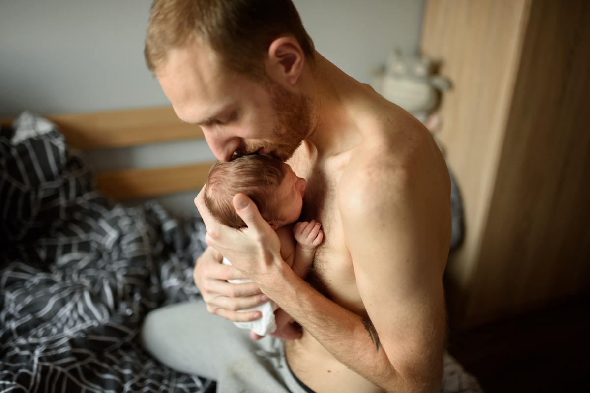 Papai e recém-nascido realizando o método canguru no quarto de casa. Ambos são brancos. Pai beija testa do bebê