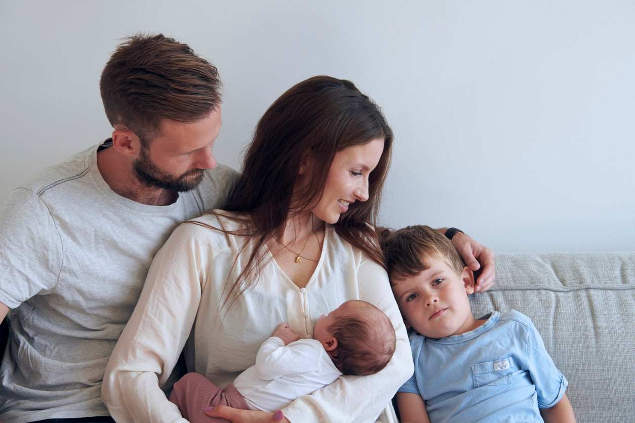 Família abraçada no sofá. O pai envolve a mãe e o filho maior, e no meio está a mãe com o bebê no colo.