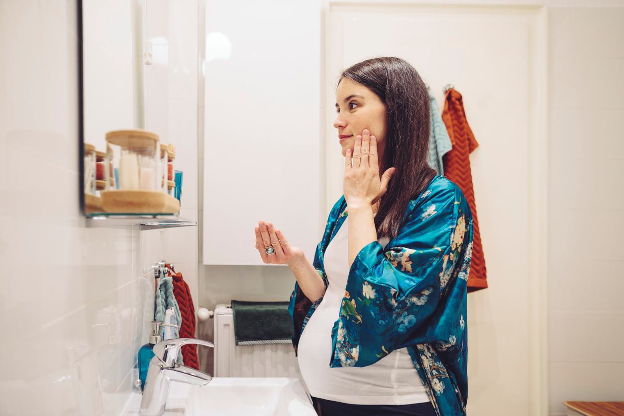 Mulher grávida fazendo skincare enquanto se olha no espelho do banheiro.