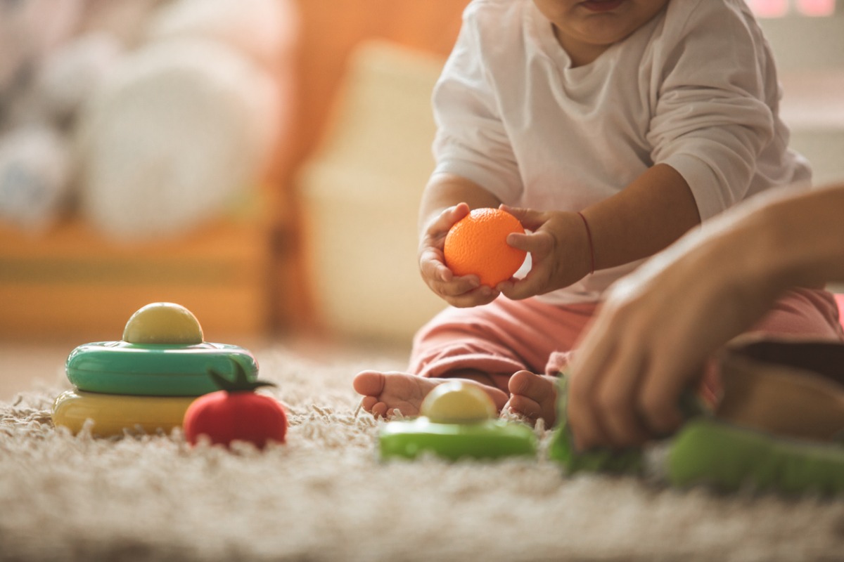 Bebê de pele clara brinca no chão, sobre tapete, com brinquedos de cores e formas diferentes. Em artigo sobre visão do bebê 