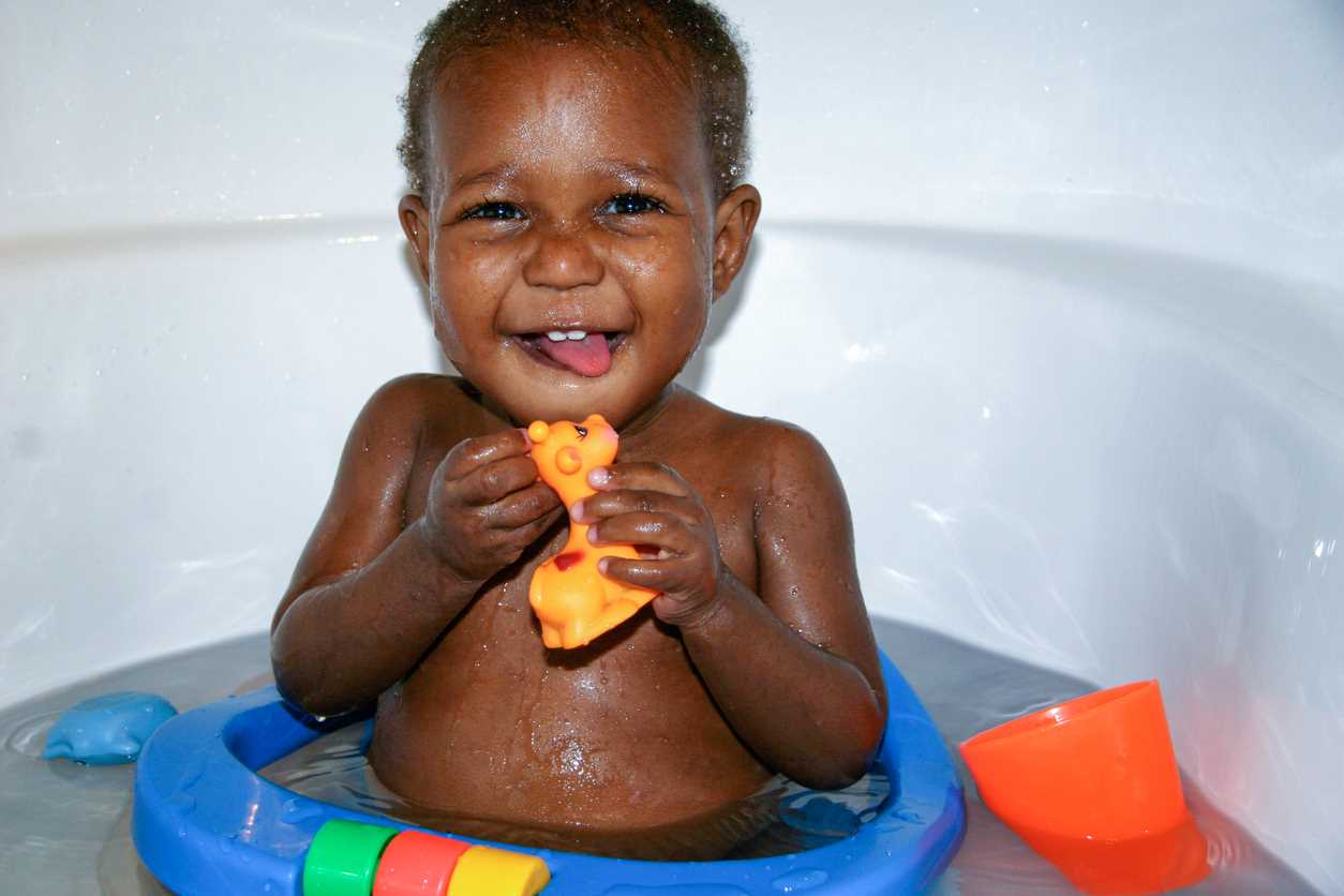 Bebê sentado na banheira, sorrindo, com brinquedos na água e segurando girafinha de borracha.