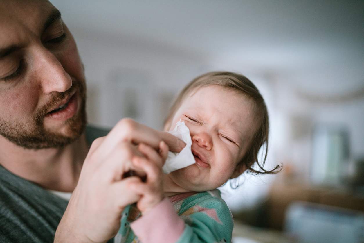 pai limpando o rosto de seu bebê com um lenço enquanto o bebê chora. O pai segura delicadamente o bebê com uma mão e usa a outra mão para limpar o rosto do bebê. 