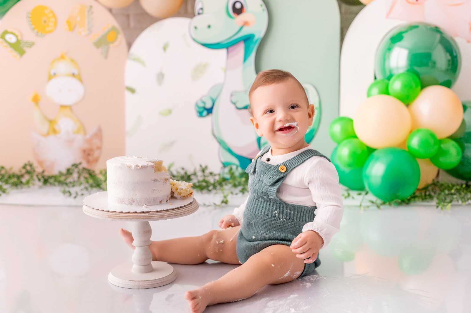 Bebê usando macacão azul está com um "bigodinho" de chantilly após ter provado do bolo que está à sua frente.