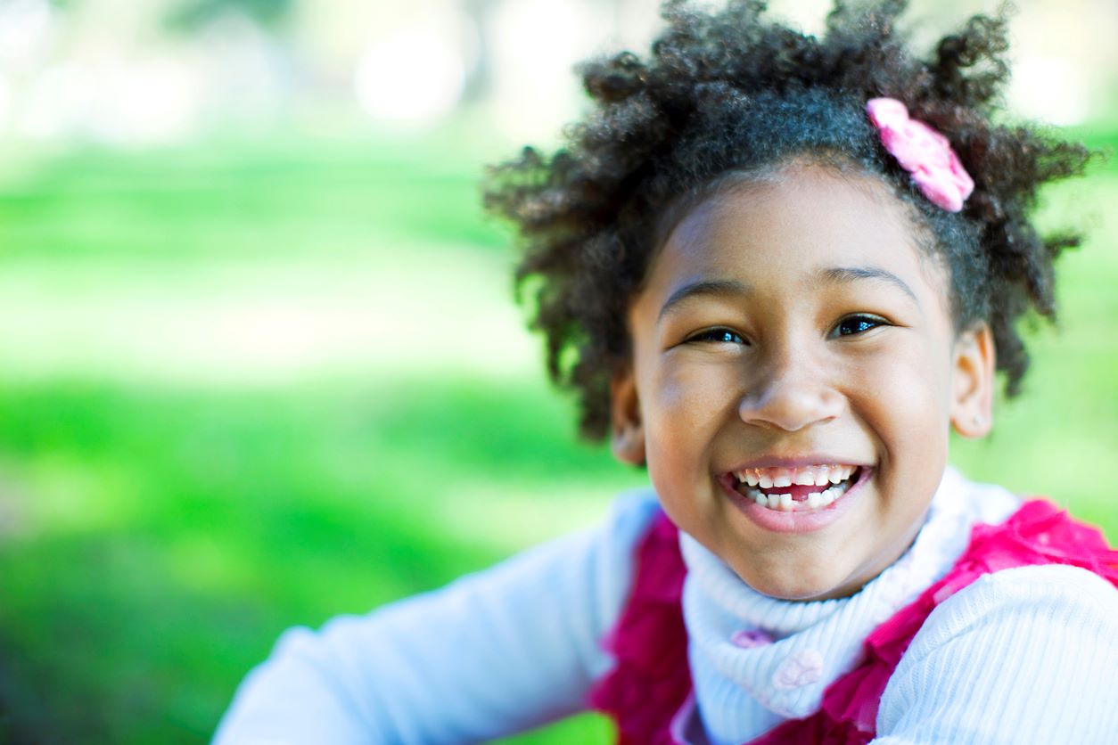 Menina sorridente no parque, feliz com penteado composto de presilha rosa no cabelo crespo