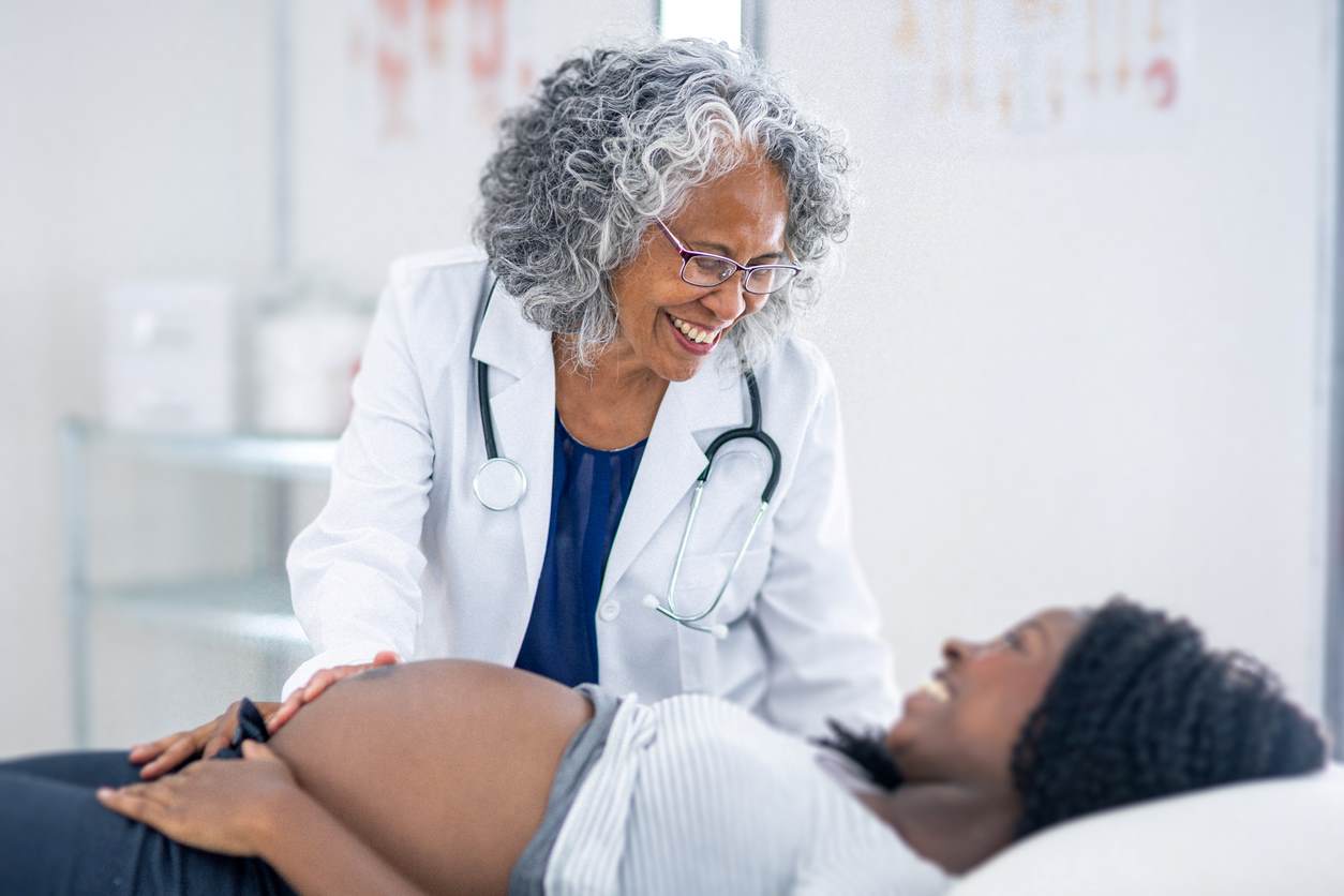 uma médica sorridente examinando a barriga de uma mulher grávida deitada em uma mesa de exame.