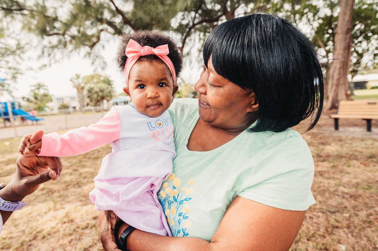 Bebê no colo da mãe, com faixa rosa no cabelo crespo, aproveitando o dia no parque