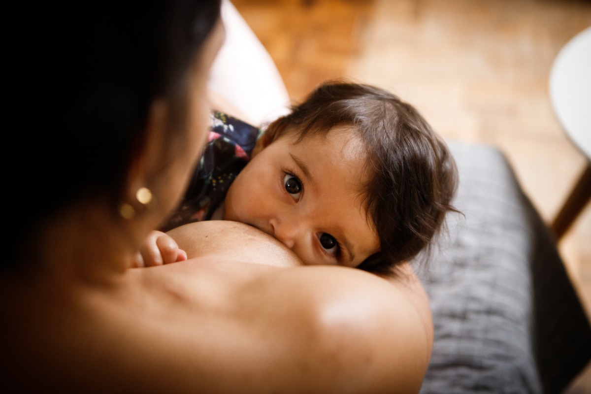 Imagem de um bebê amamentando no peito da mãe. O bebê tem cabelos escuros e está olhando para a câmera enquanto mama.