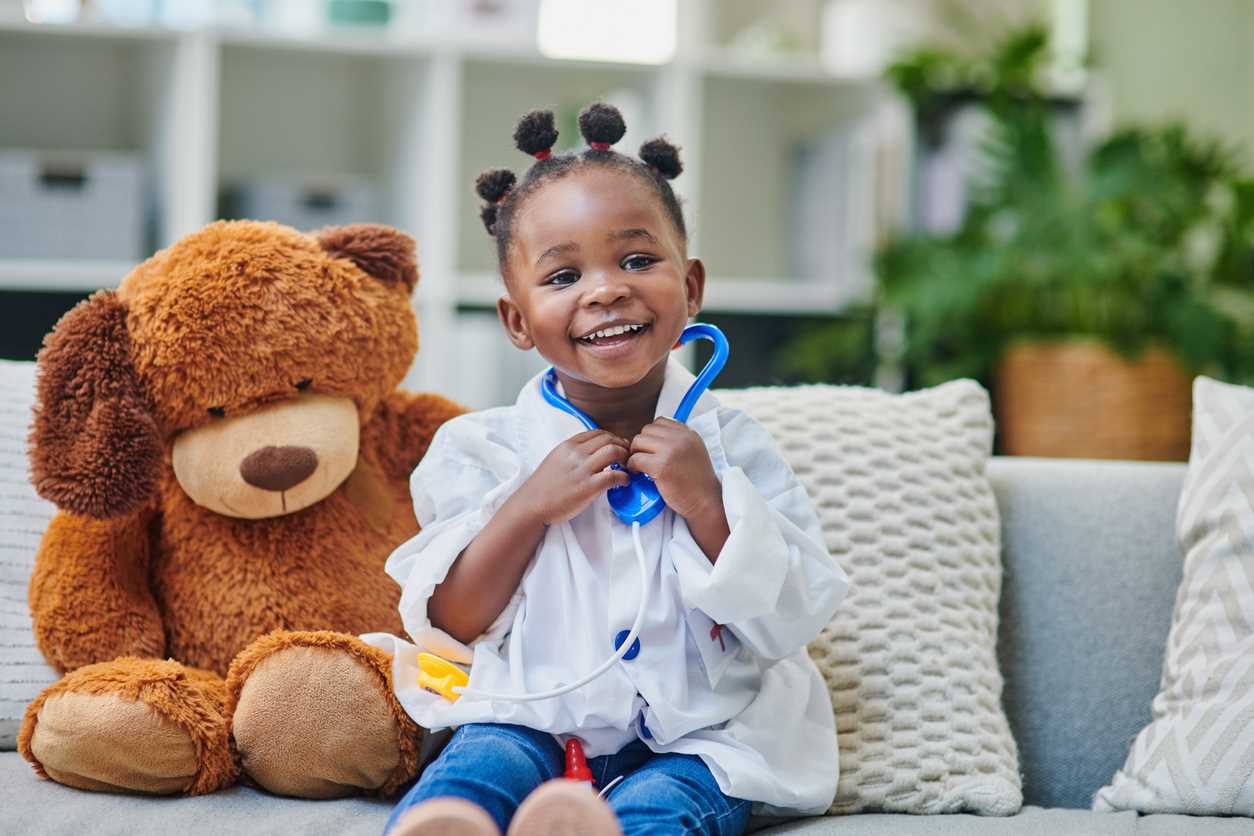 Menina pequena sentada no sofá de casa, vestida de médica ao lado de pelúcia, feliz com seu penteado afro puffs