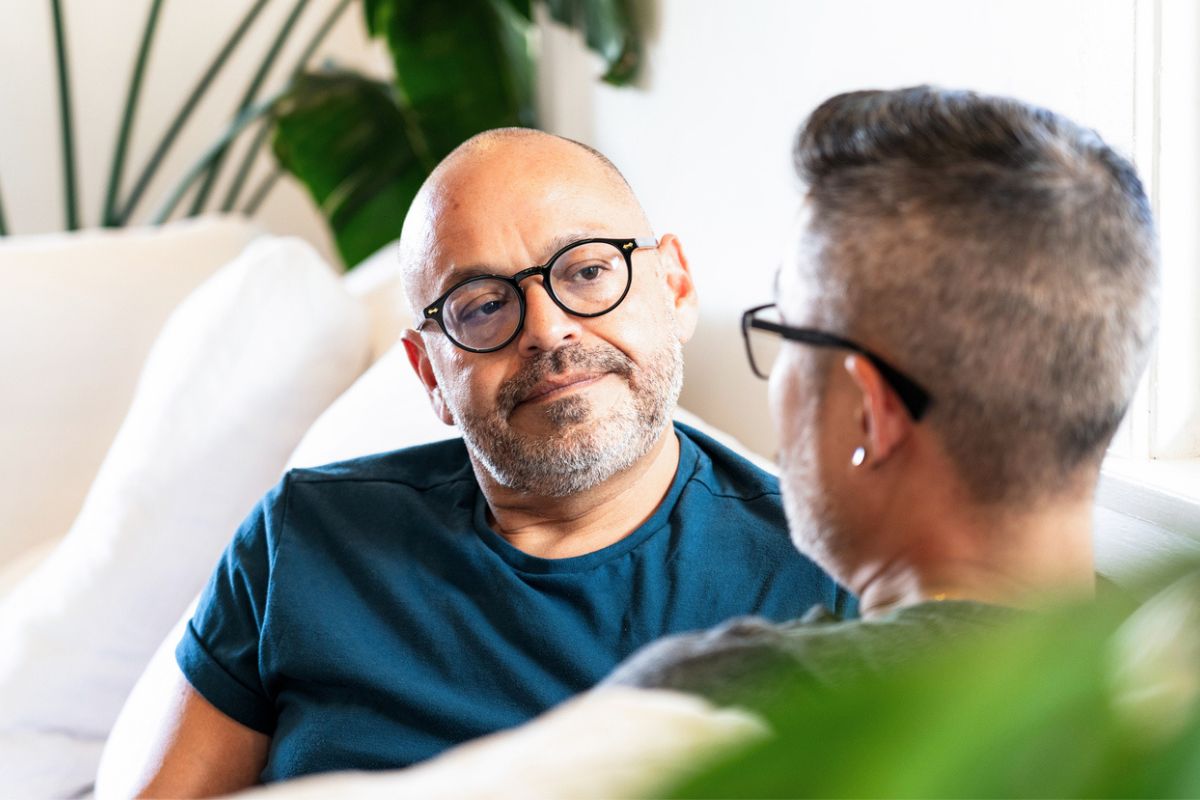 casas de dois homens sentados em um sofá, tendo uma conversa. Ambos usam óculos e um deles, com barba e cabeça raspada, está olhando atentamente para o outro.