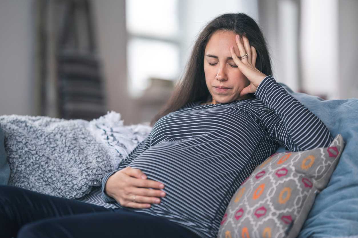 Gestante sentada em sofá, com uma mão apoiando o rosto e outra a barriga, com os olhos fechados.