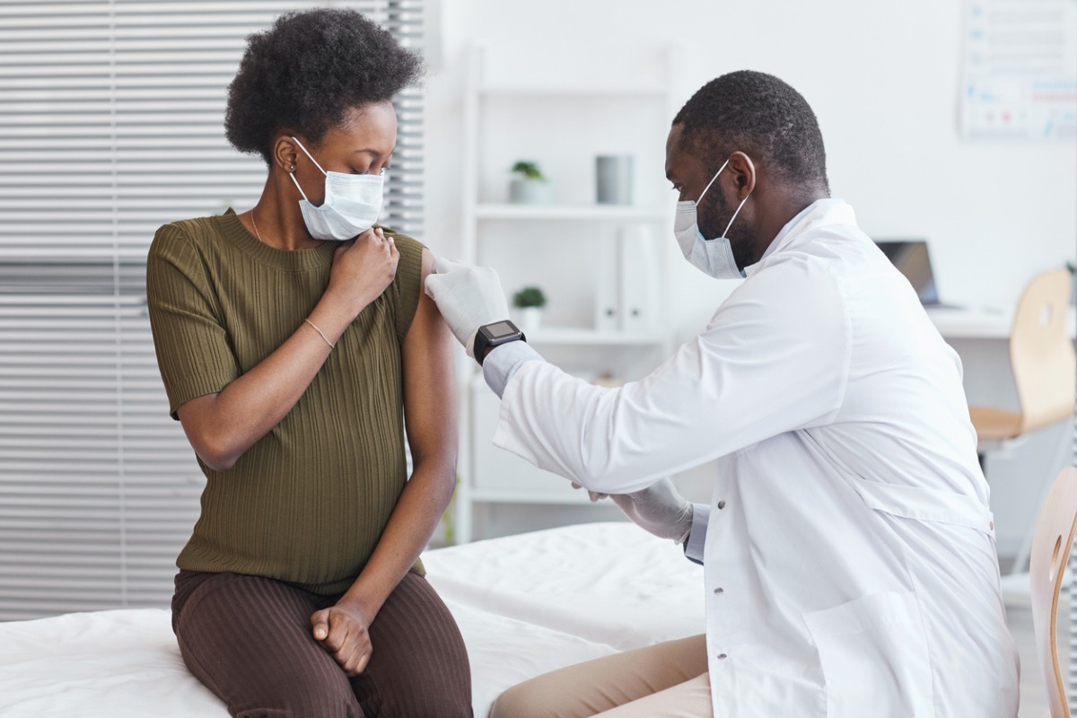 Mulher negra recebendo vacina na gestação. O médico, também negro, é quem aplica a vacina, no consultório médico. 