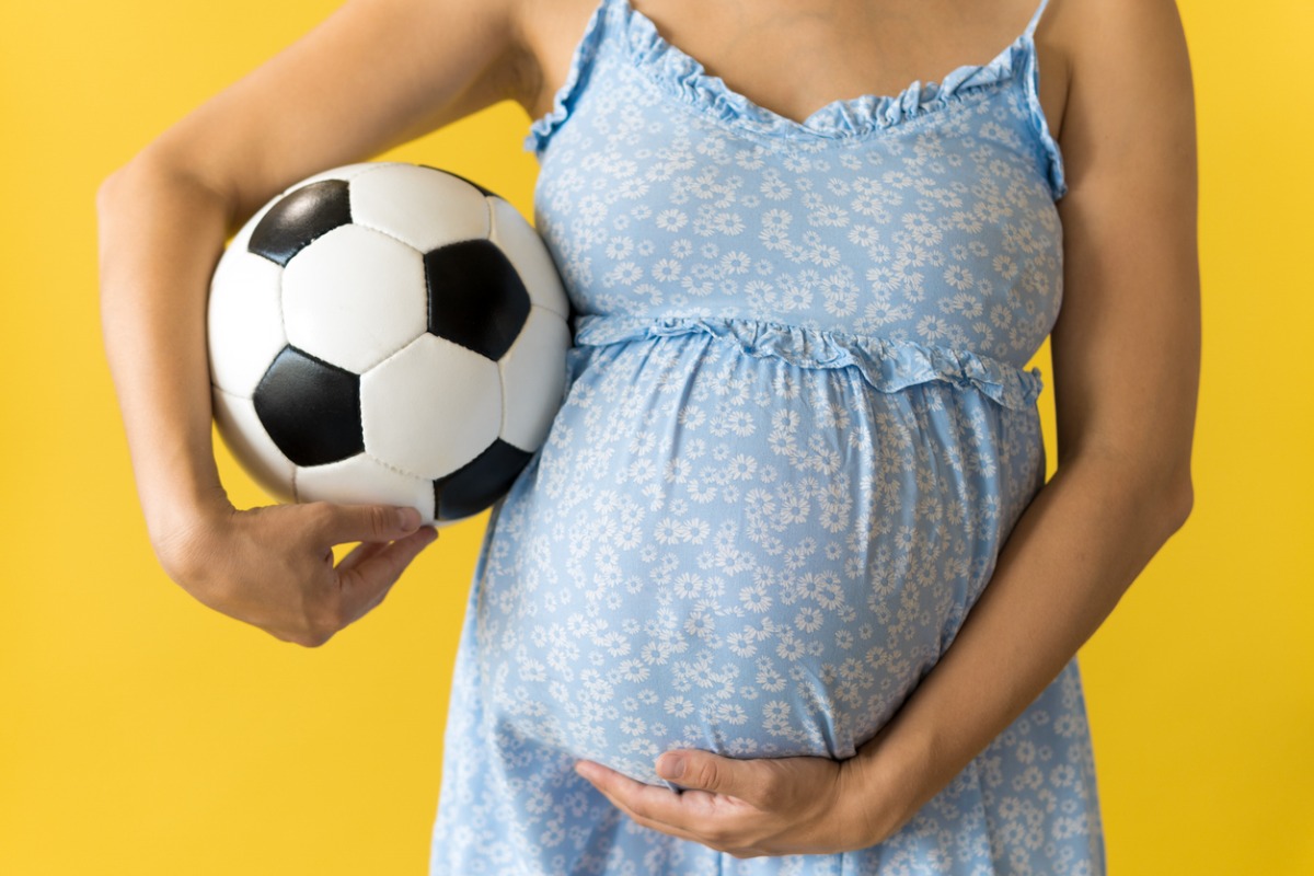 Imagem de uma mulher grávida vestindo um vestido azul com estampas florais, segurando uma bola de futebol debaixo do braço.