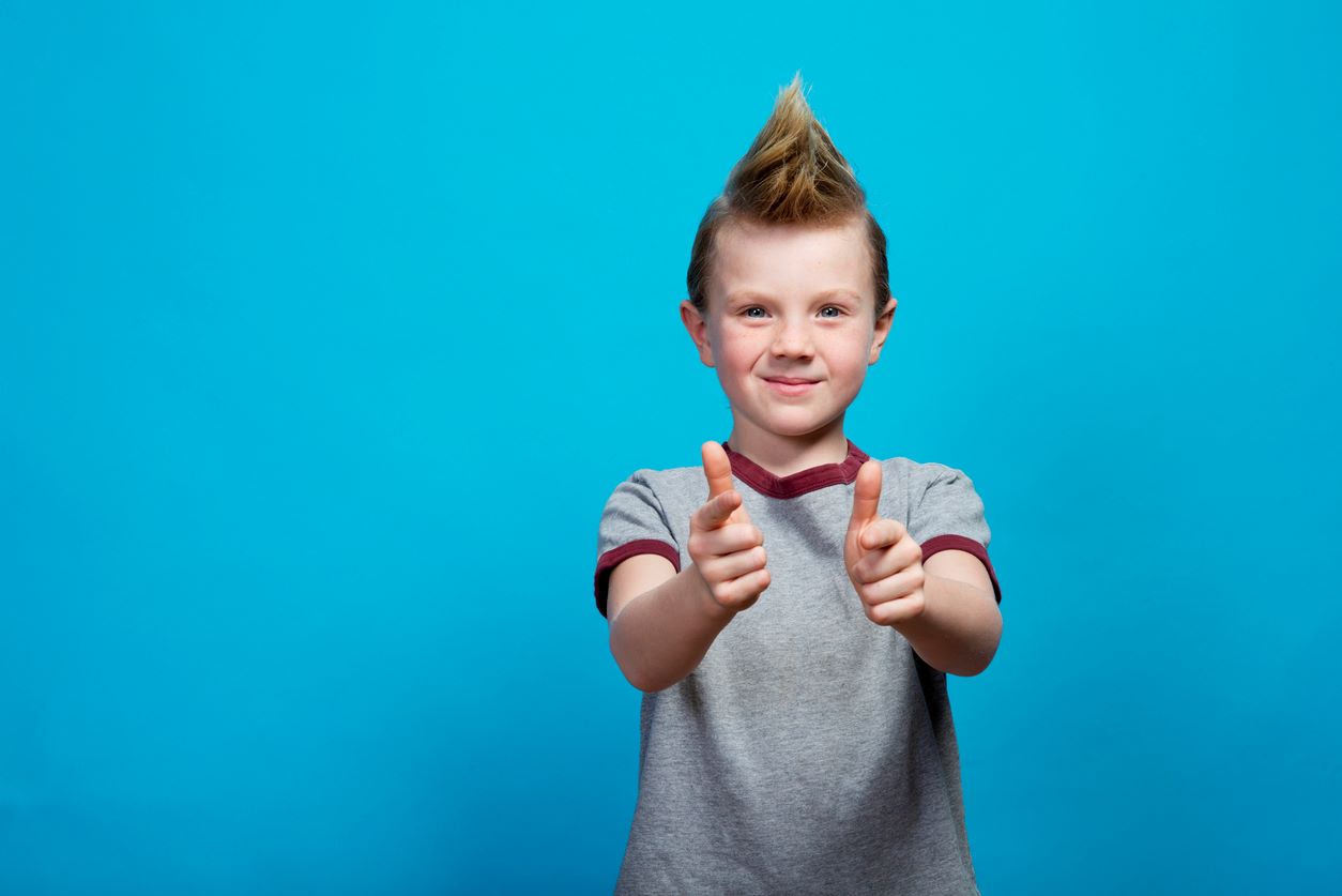  Menino com cabelo estilo moicano, fazendo sinal de positivo com as duas mãos e sorrindo para a foto.