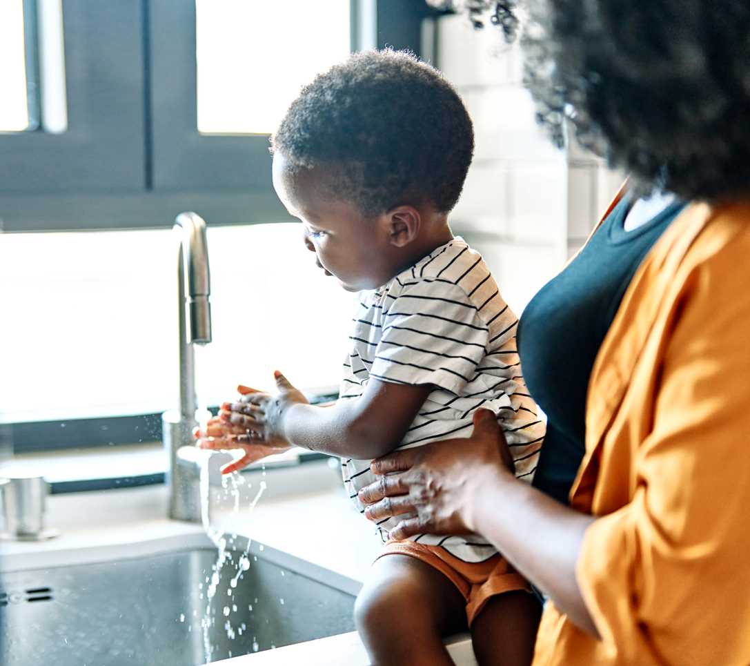 mãe ajudando seu filho pequeno a lavar as mãos na pia da cozinha. A mãe está segurando o filho, que está sentado na beirada da pia, enquanto a água corre sobre as mãos do menino.