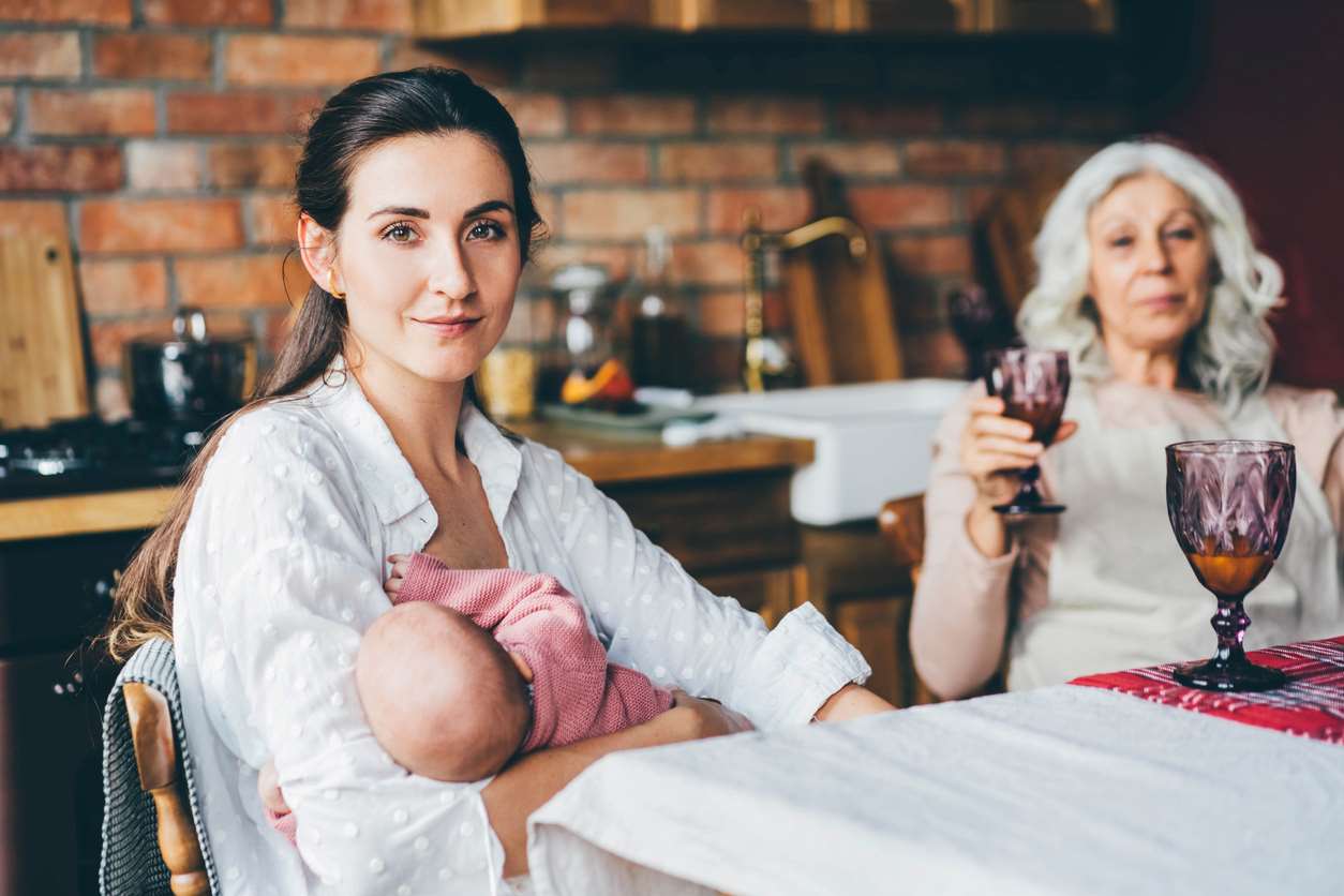 Mulher amamentando bebê  enquanto outra mulher, está ao fundo segurando uma bebida.