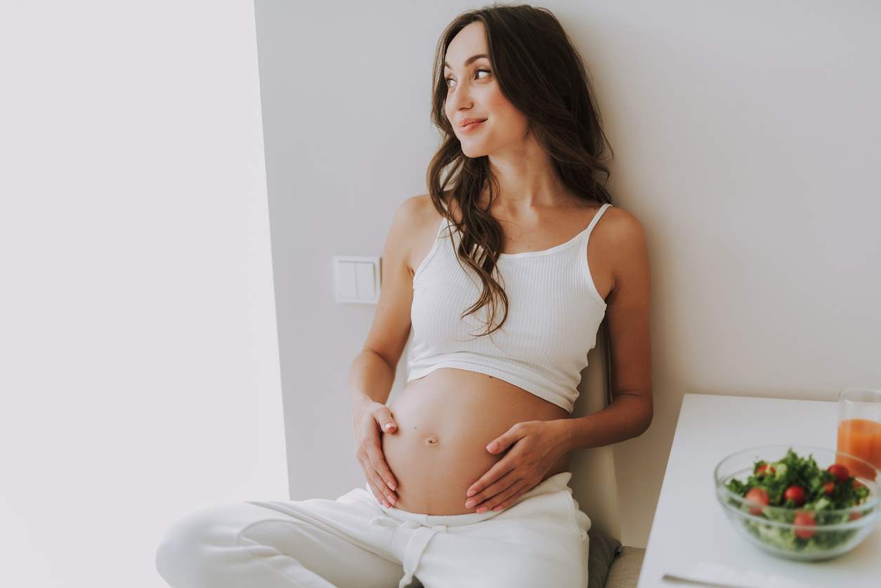 Mulher grávida, sentada, de cabelo longo, tocando a barriga e olhando para o lado.