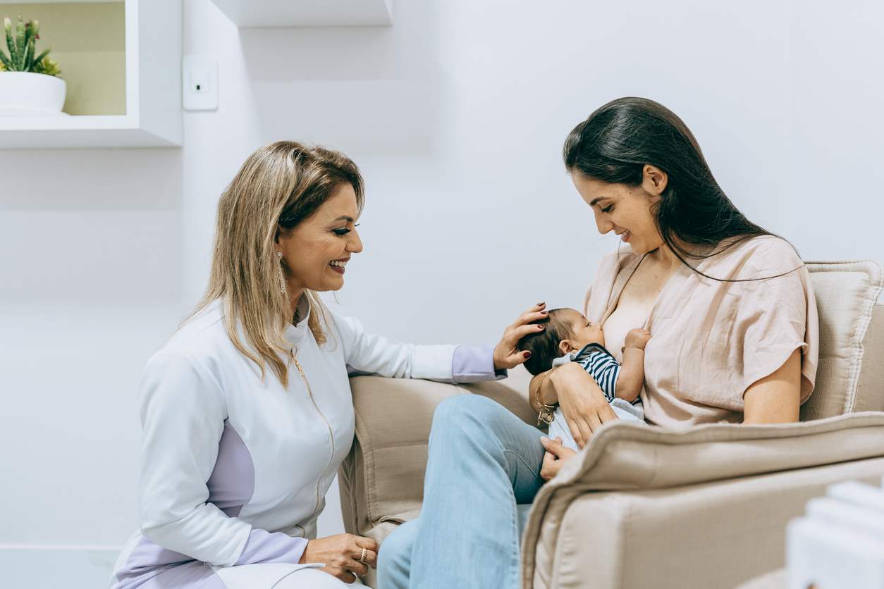 Mãe amamentando seu bebê enquanto recebe orientação de uma profissional em consultório.