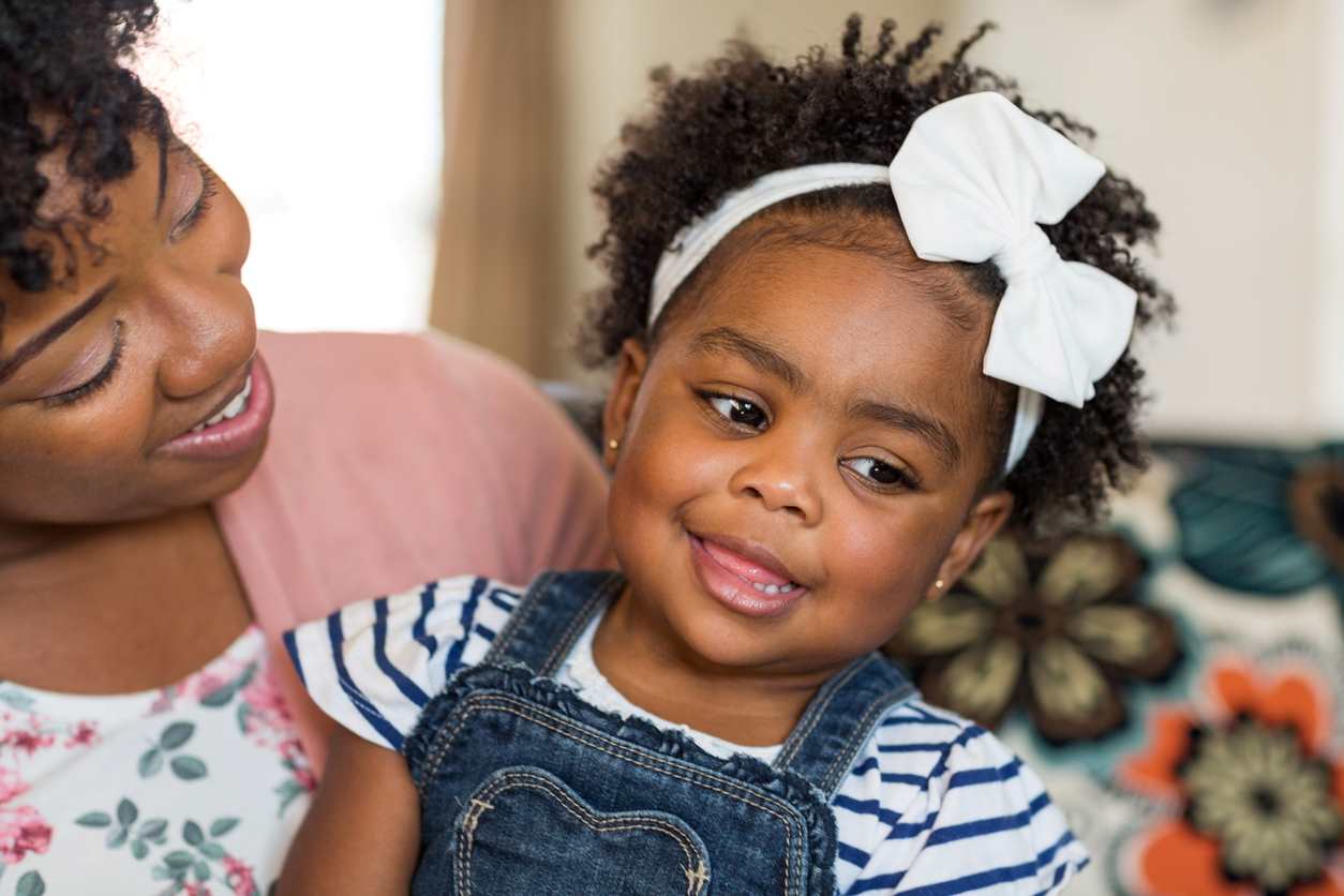 Criança pequena no colo da mãe, com penteado composto de laço branco no cabelo 