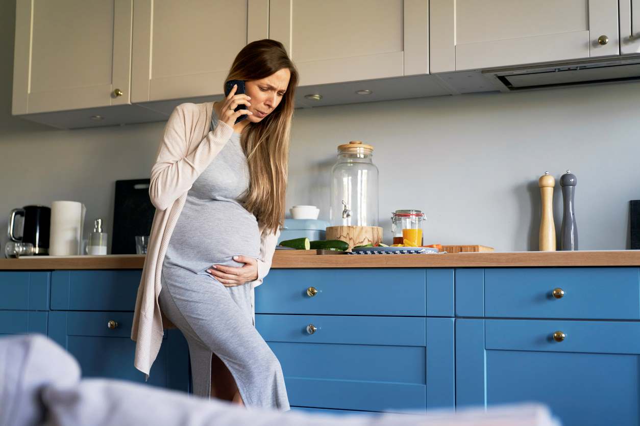 Mulher grávida ao celular, com uma mão no pé da barriga e fisionomia de dor