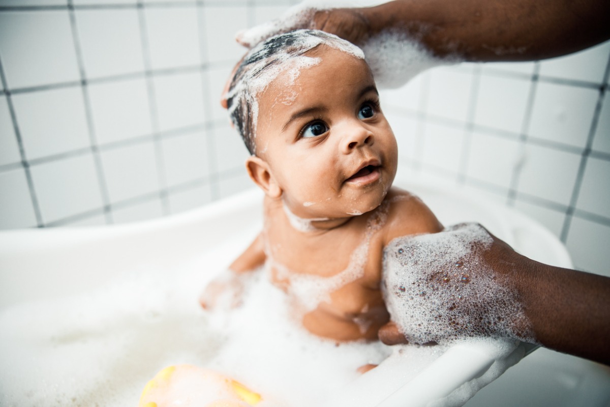 Bebê negro em banho de banheira com espuma de shampoo de bebê na cabeça. Ele para o pai que dá banho, de quem só vemos as mãos 