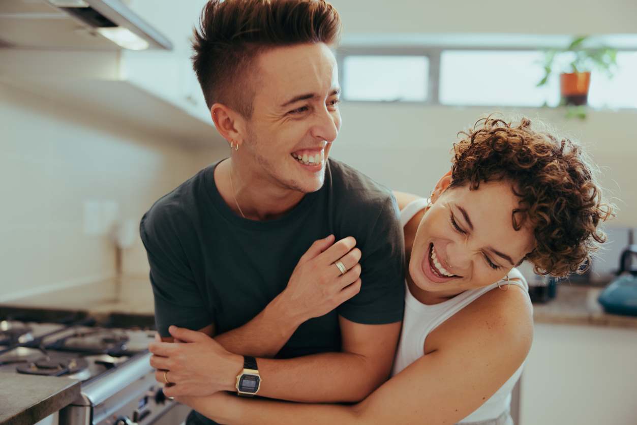 Casal LGBTQIA+ sorrindo e abraçando na cozinha.