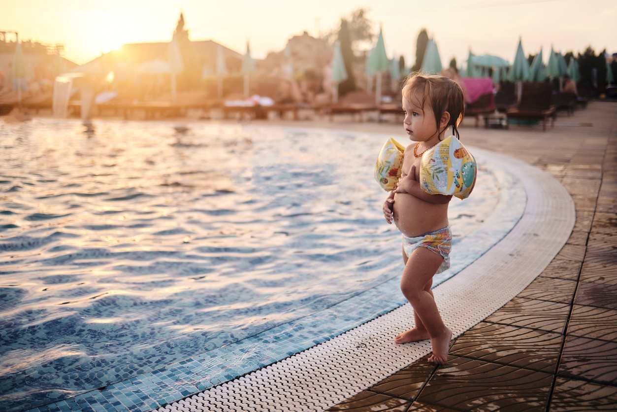 Criança com boias de braço e fralda na beira de uma piscina.